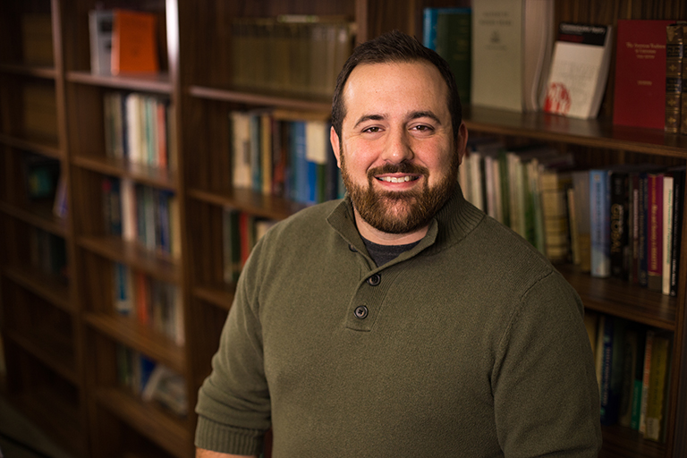 Dustin Carnahan wearing olive green sweater and standing next to bookcase.
